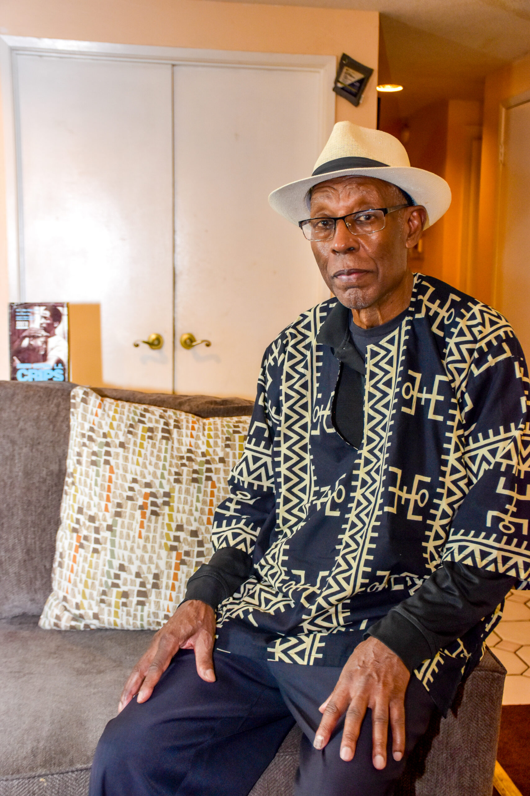 Donald Bakeer seated beside a sofa wearing a white hat and a black and gold patterned shirt