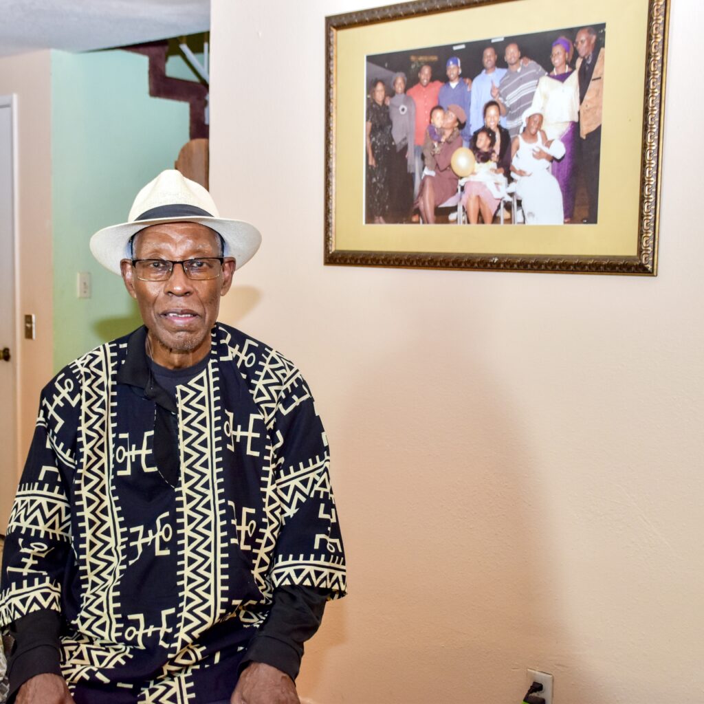 Donald Bakeer seated beneath a framed family photograph wearing a white hat and patterned shirt